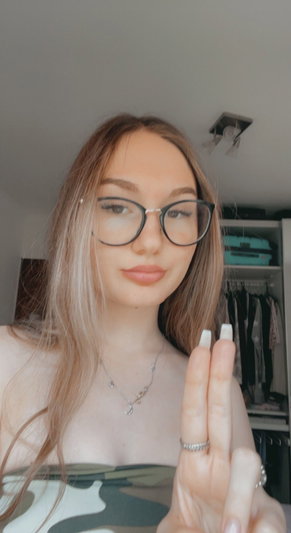 Woman with glasses and long hair in a camo top, making a peace sign indoors.