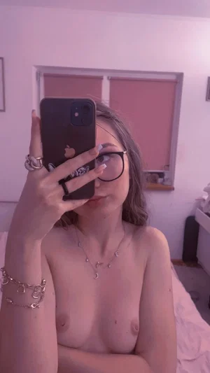 Woman with glasses and jewelry takes mirror selfie, bare-chested, in pink-lit room.