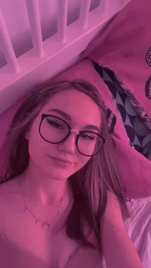 Woman with glasses lying in bed, wearing a thin top, pink lighting, indoor setting.