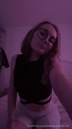 Woman with glasses in black crop top and white shorts sitting on bed in dim purple light.