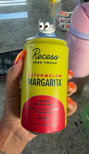 Hand with bright orange nails holding a yellow can of Recess watermelon margarita mocktail.