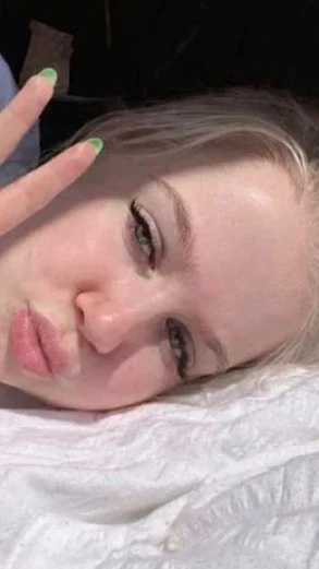 Woman with blonde hair and green nail polish lying on white sheets, making a playful face.