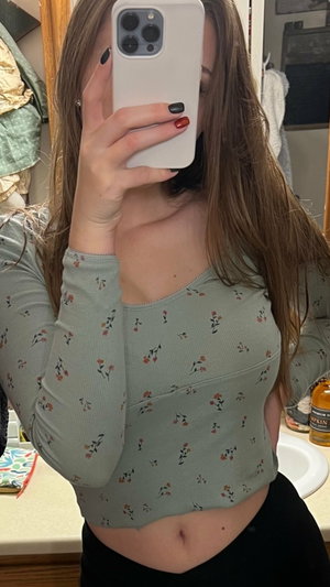 Woman in floral crop top and black pants taking mirror selfie in bathroom.