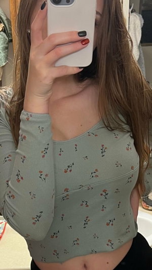 Woman with long brown hair in floral crop top, holding phone for mirror selfie.