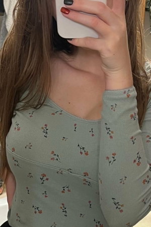 Woman with long brown hair in floral top taking mirror selfie indoors.