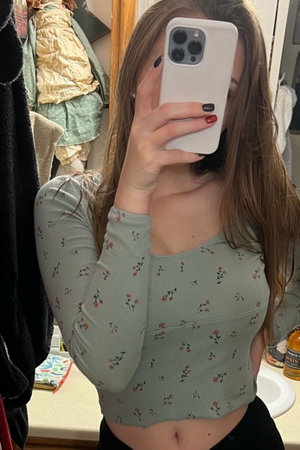 Woman in floral top and black bottoms takes mirror selfie in bathroom.