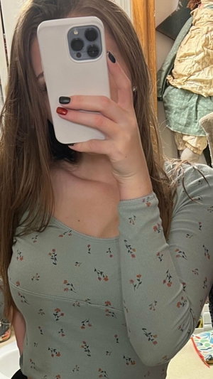 Woman with long brown hair in floral top taking mirror selfie indoors.