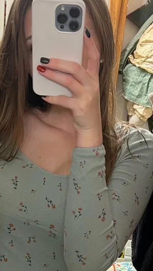 Woman with long brown hair in floral top taking mirror selfie indoors.