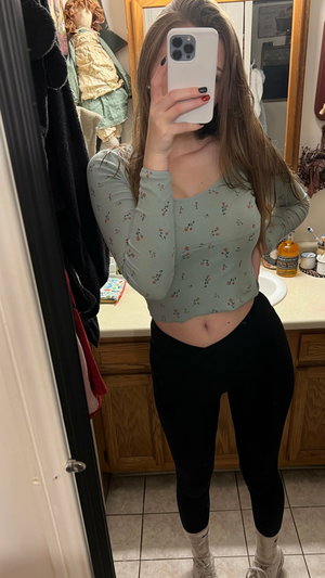 Woman in floral crop top and black leggings taking mirror selfie in bathroom.