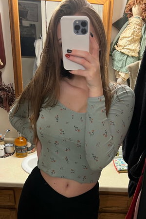 Woman with long brown hair in floral crop top and black pants, taking mirror selfie in bathroom.
