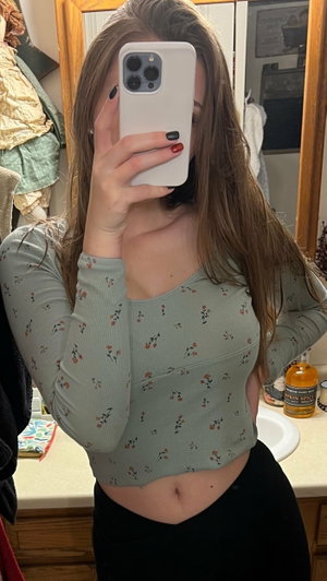 Woman with long brown hair in floral crop top and black pants, taking mirror selfie in bathroom.