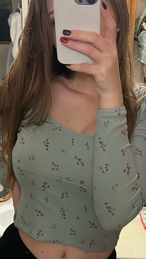 Woman with long brown hair in floral top taking mirror selfie in bedroom.