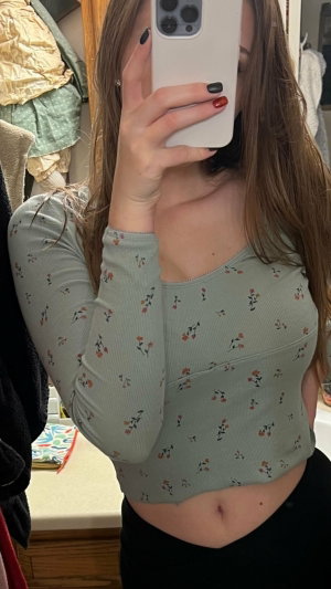 Woman in floral top and black bottoms takes mirror selfie in bedroom.