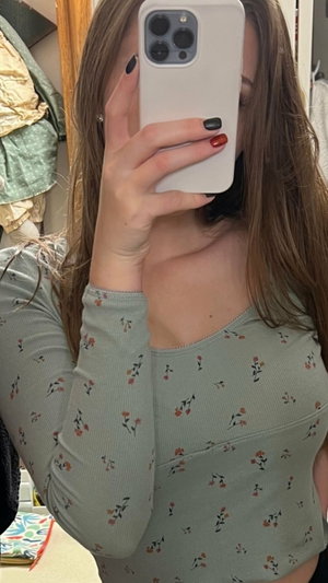 Woman with long brown hair in floral top taking mirror selfie indoors.
