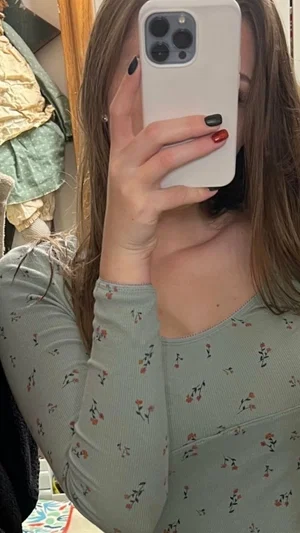 Woman with long brown hair in floral top taking mirror selfie indoors.