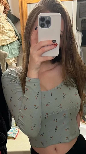 Woman with long brown hair in floral top taking mirror selfie in bedroom.