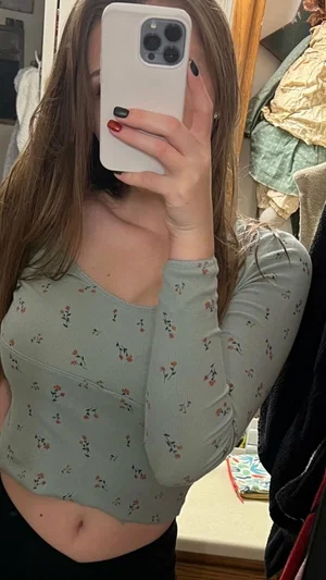 Woman in floral crop top and black pants taking mirror selfie in bedroom.