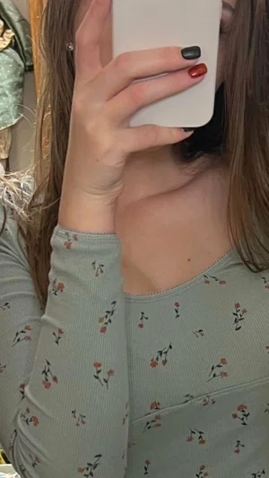 Woman in floral long-sleeve top taking mirror selfie, face obscured by phone.