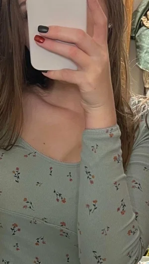 Woman with long brown hair in floral long-sleeve top taking mirror selfie indoors.