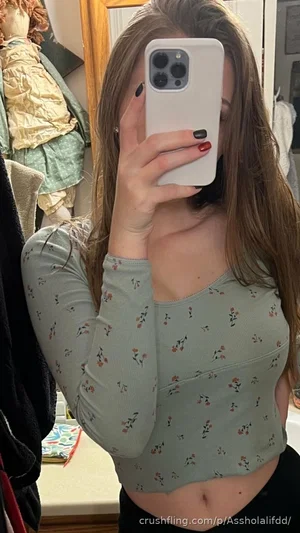 Woman in floral crop top and black pants, taking mirror selfie in bedroom.