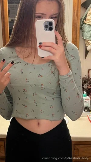 Woman in floral crop top and black skirt taking mirror selfie in bathroom.