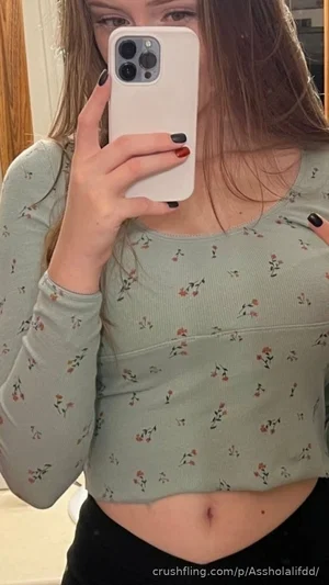 Woman in floral crop top and black pants taking mirror selfie, midriff exposed.