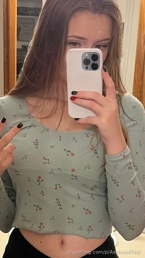 Woman in floral crop top and black pants taking mirror selfie, midriff exposed.