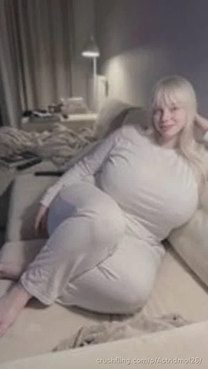 Blonde woman in white loungewear sitting on a bed in a dimly lit bedroom.