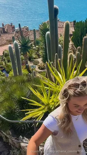 Blonde woman in white top and vest, outdoors near cacti and ocean.