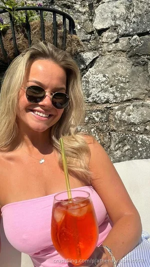 Woman with blonde hair in pink strapless top holding drink outdoors near stone wall.