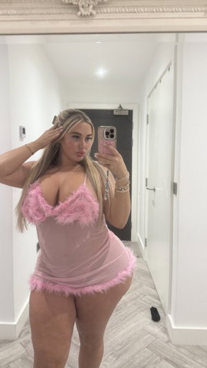 Blonde woman in pink feathered dress taking mirror selfie indoors.