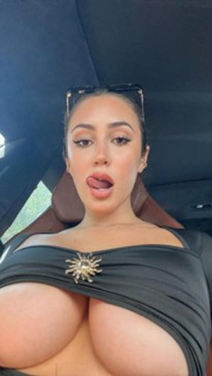 Woman in black off-shoulder top with sun brooch, sitting in car, tongue out.