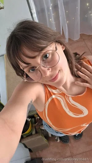 Woman with glasses and brown hair in orange tank top, taking selfie indoors.