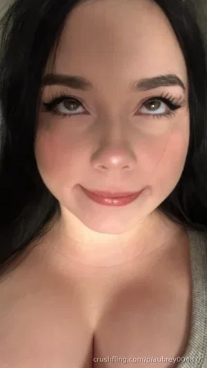 Woman with dark hair and makeup, wearing a gray top, smiling in a close-up selfie.