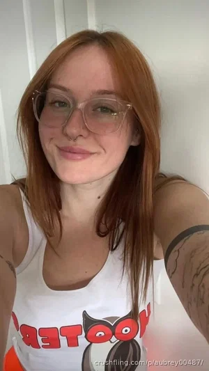 Woman with red hair and glasses wearing a white tank top, indoors.