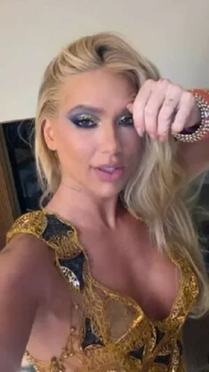 Blonde woman in gold lace top with black mesh, posing indoors.