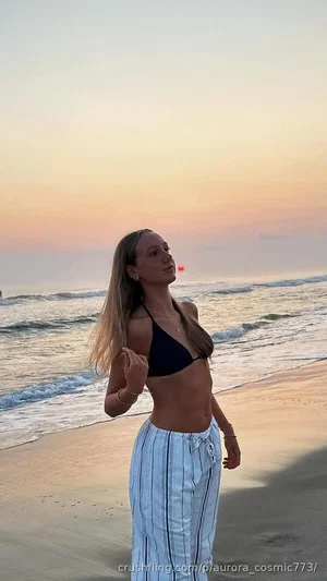Woman in black bikini top and striped pants standing on beach at sunset.