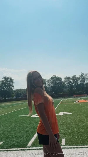 Woman with long blonde hair in orange t-shirt and black shorts on a football field.