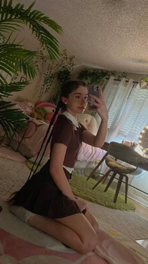 Woman in schoolgirl outfit kneeling on bed in bedroom with plants and soft lighting.