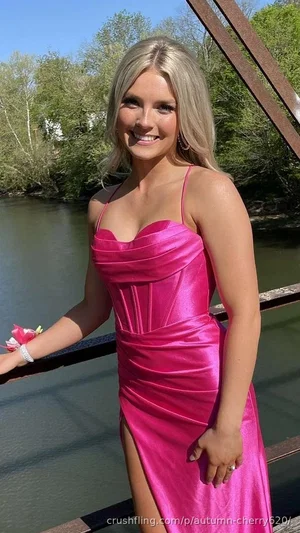 Woman in pink dress standing by river, blonde hair, smiling.