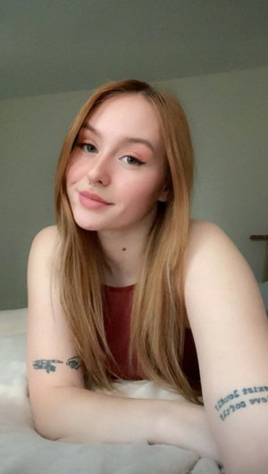 Woman with long blonde hair and tattoos lying on bed, wearing red top.