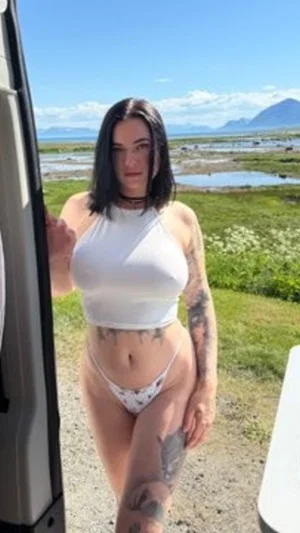Woman with tattoos in white crop top and patterned underwear standing outdoors near a vehicle.