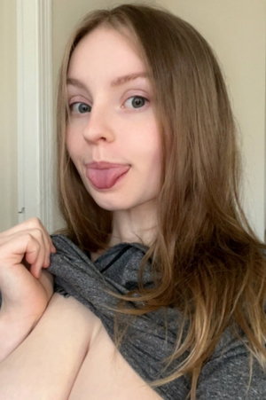 Woman with brown hair sticks tongue out, pulls gray top down indoors.
