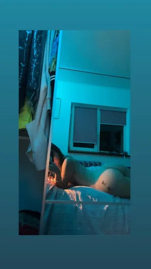 Dark-haired woman lying nude on bed taking mirror selfie.