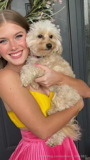 Woman in yellow top and pink skirt holding a small dog, smiling in front of a door with flowers.