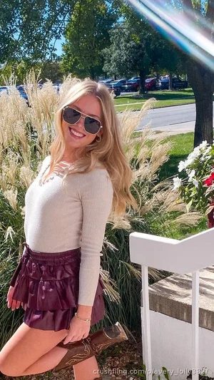 Blonde woman in beige top and purple skirt posing outdoors near plants and flowers.