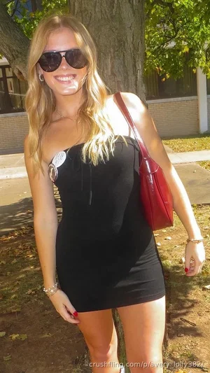 Woman in black strapless dress, sunglasses, red purse, standing outdoors near tree.