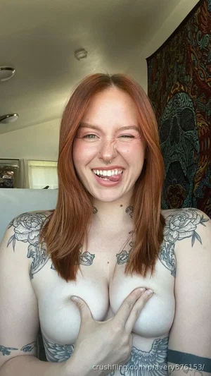 Woman with red hair and tattoos covering breasts with hands, indoors.