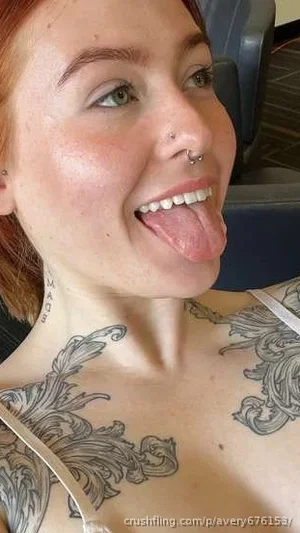 Woman with red hair and tattoos on chest, wearing white top, sticking out tongue indoors.