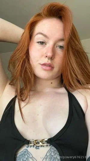 Woman with red hair in black top with gold chain detail, chest tattoo visible, indoors.